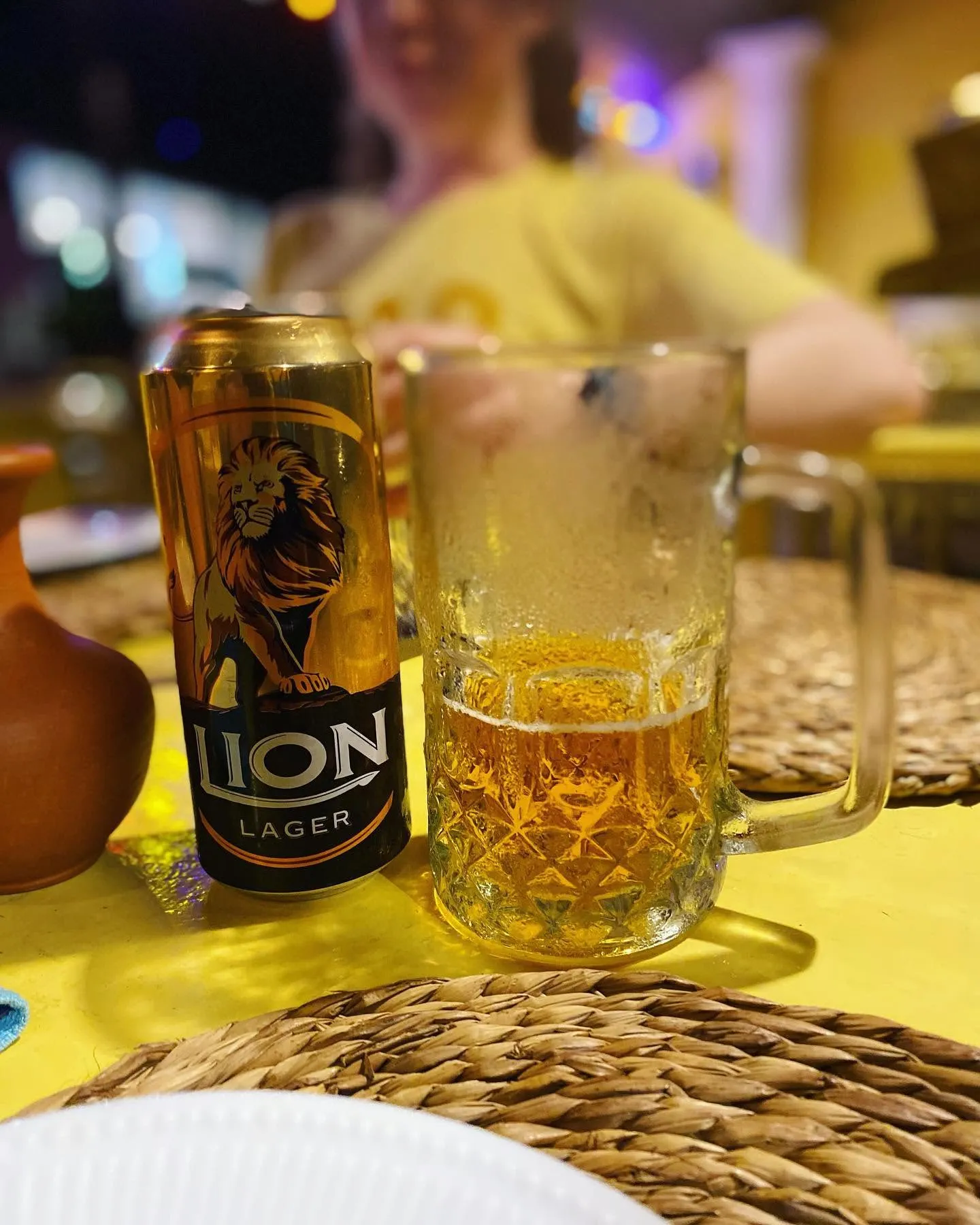 Lion Lager can and glass beer mug on a yellow tablecloth in a warmly lit restaurant